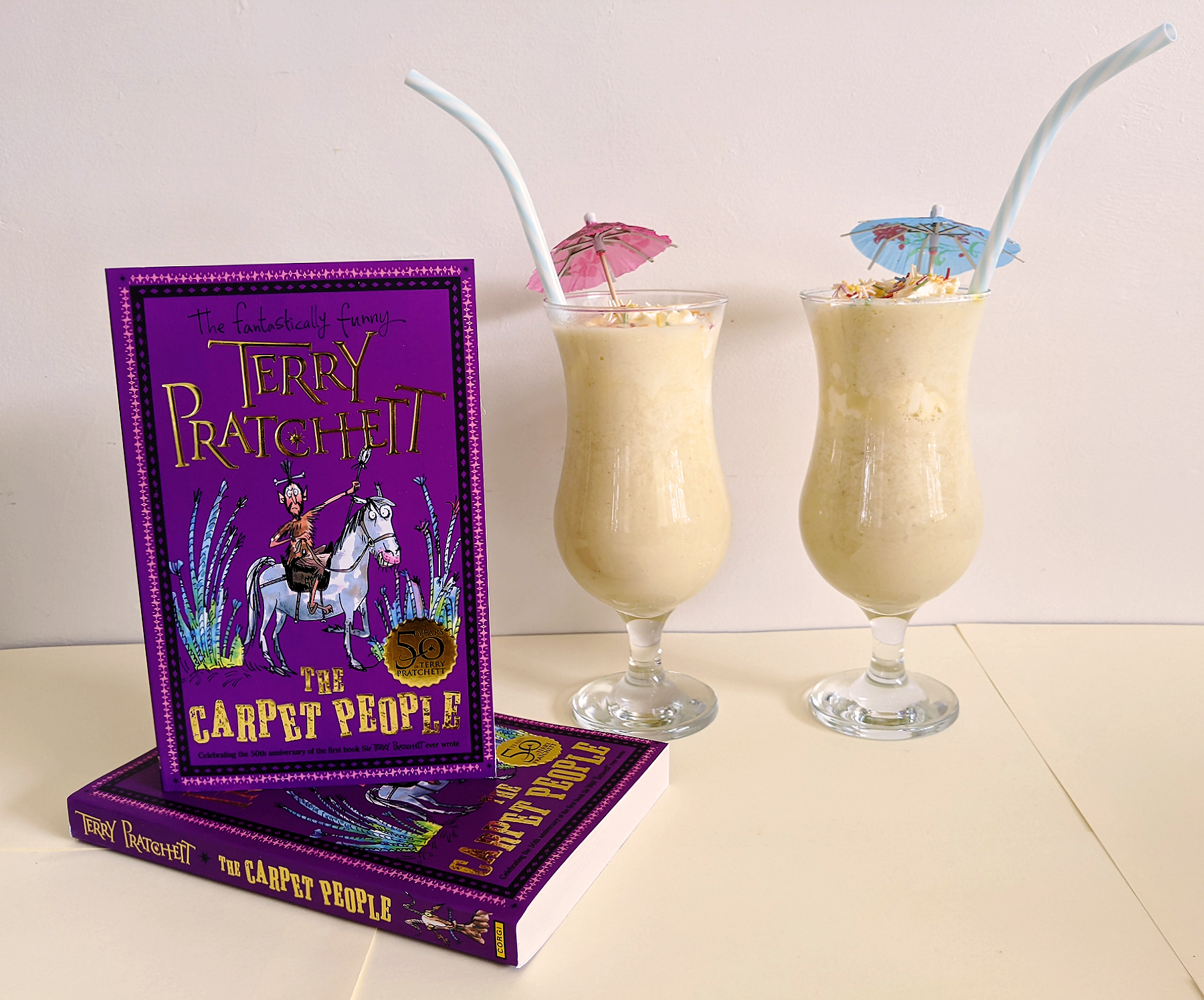 A photo of two copies of the 50th anniversary edition of The Carpet People by Terry Pratchett and two banana milkshakes in tall glasses with straws and mini umbrellas