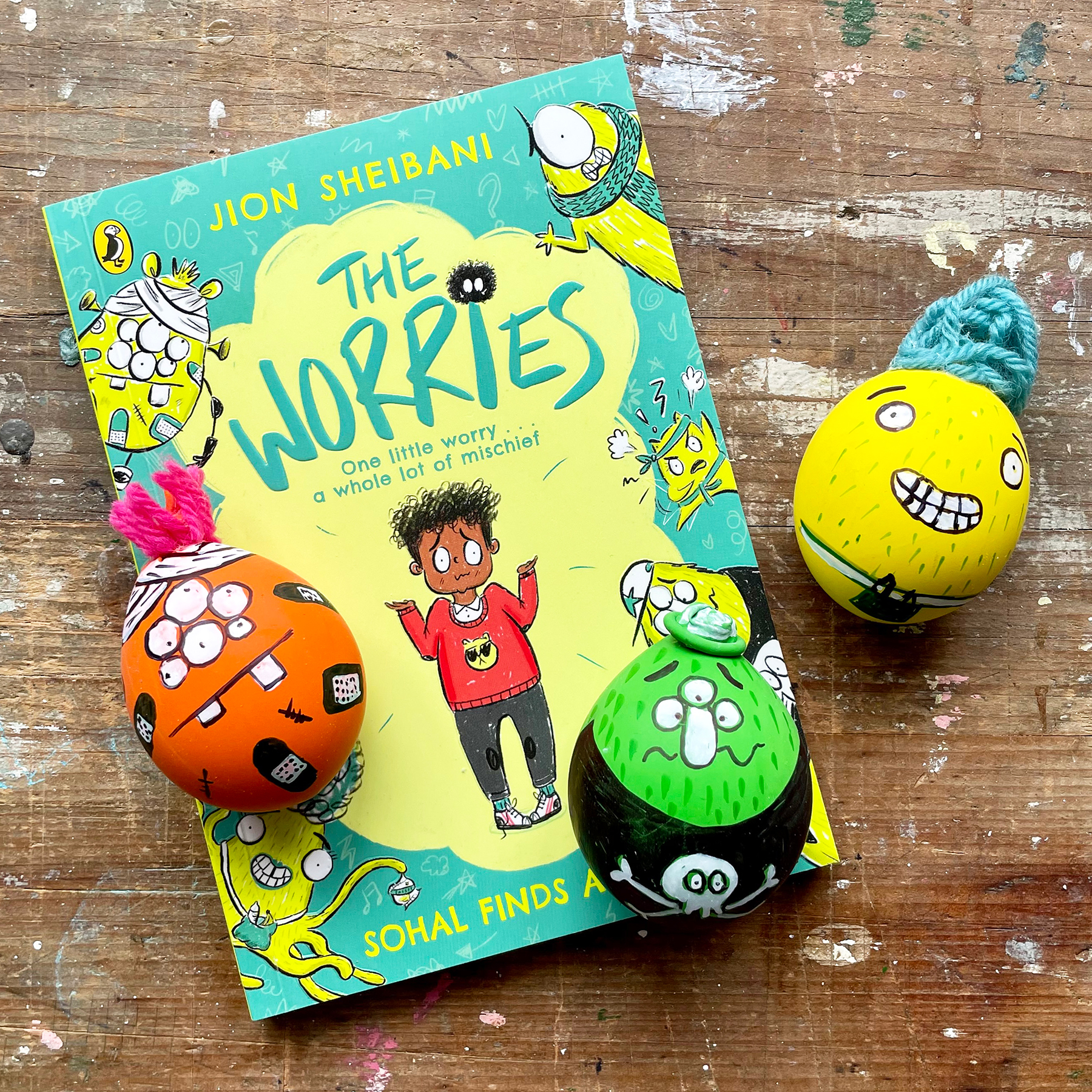 A photo of the final result, three funny face stress balls, one green, one yellow and one orange next to the book The Worries by Jion Sheibani