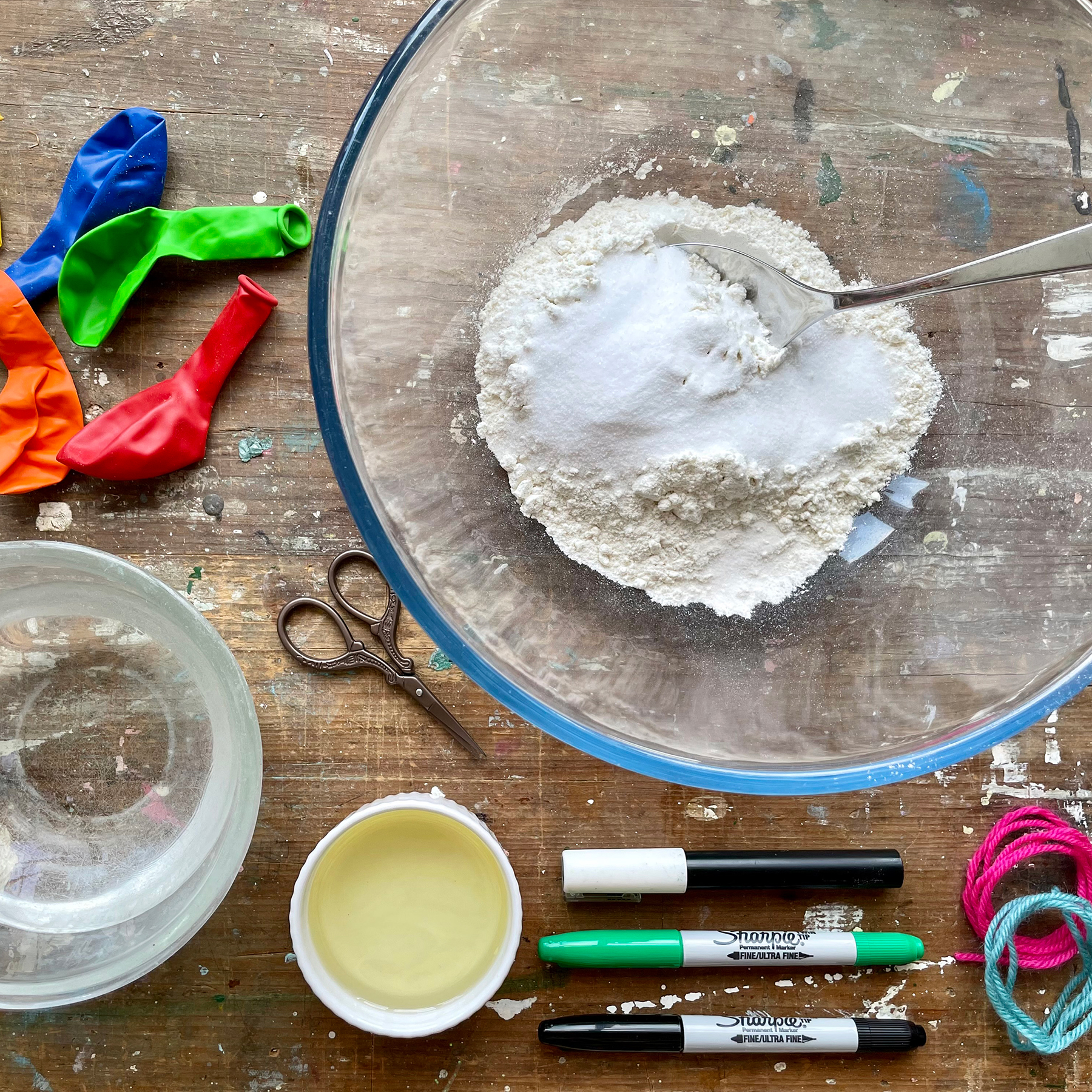A photo of all the things you will need to make a stress ball, including balloons, a bowl of plain flour and salt, warm water, oil, marker pens, wool, glue and a pair of scissors; they sit on a wooden table
