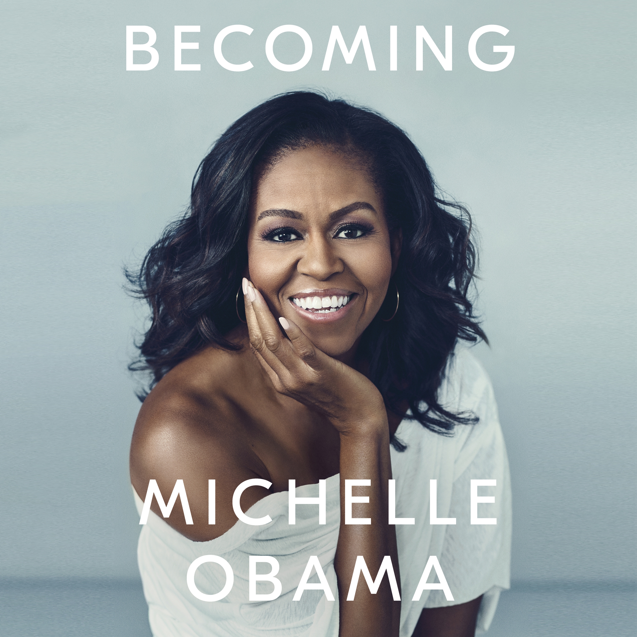 Audiobook image for Becoming: a grey background with a photograph of Michelle Obama taking up most of the cover. She's smiling to camera, resting her chin on her hand, wearing a white off-shoulder top.  The title is at the top in white font, and the author's name is at the bottom in bigger white font.