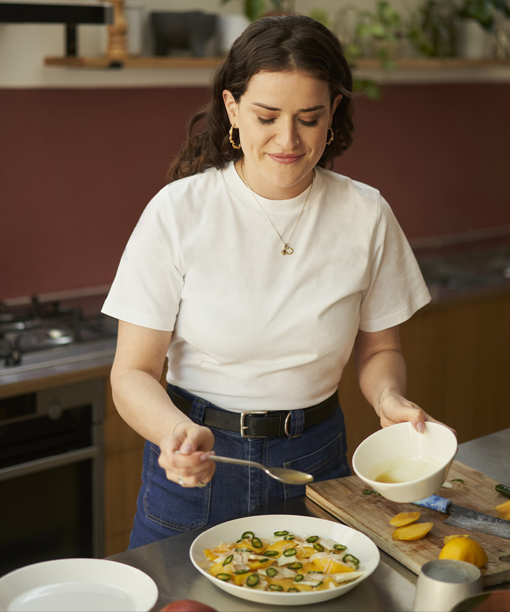 A photograph of ixta belfrage pouring dressing over a salad of oranges. She is wearing a white teeshirt.
