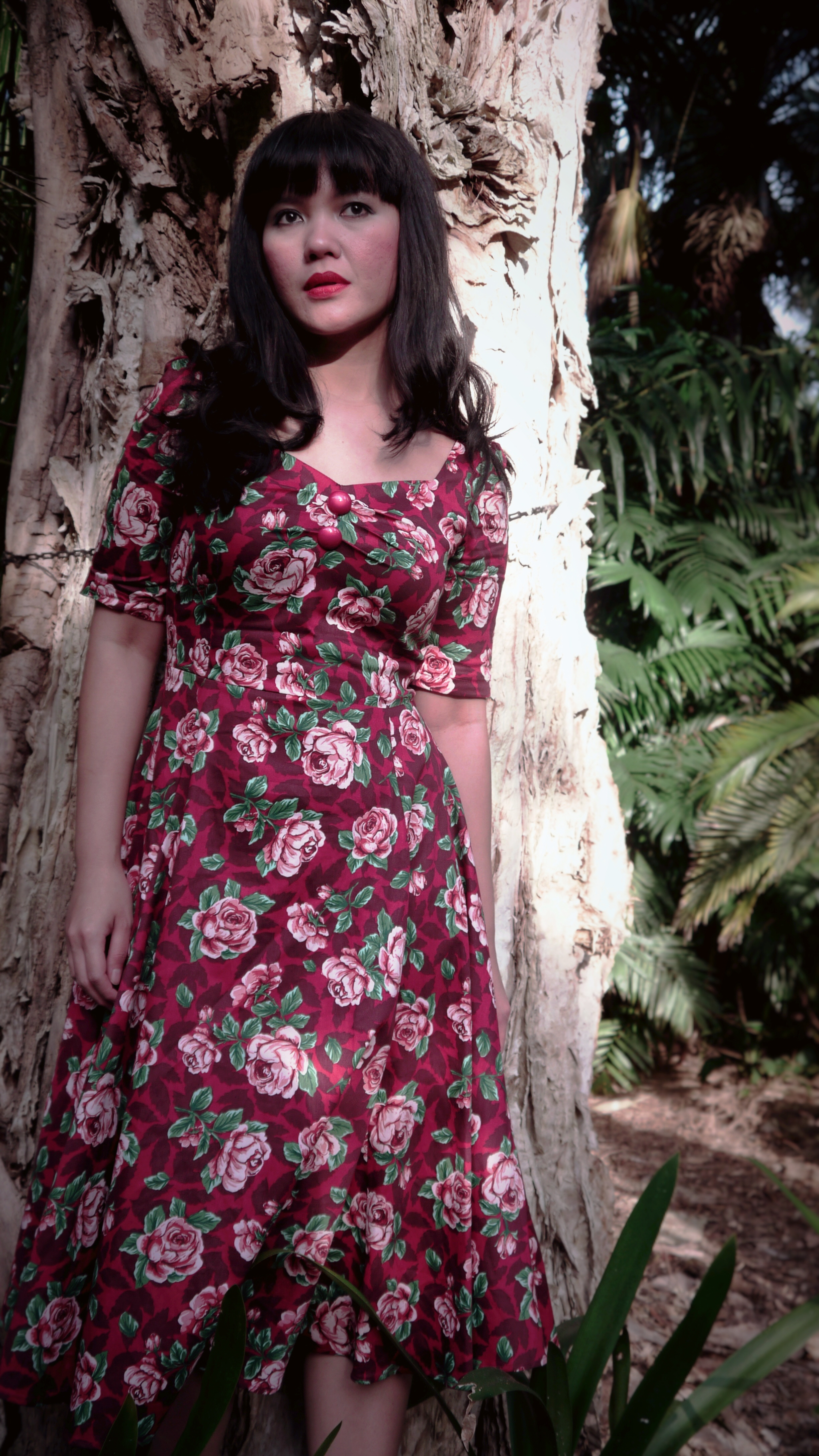 Photo of Intan Paramaditha, standing outdoors against a tree trunk. She is an Indonesian woman with long, dark hair and a fringe, wearing a knee-length red floral dress.