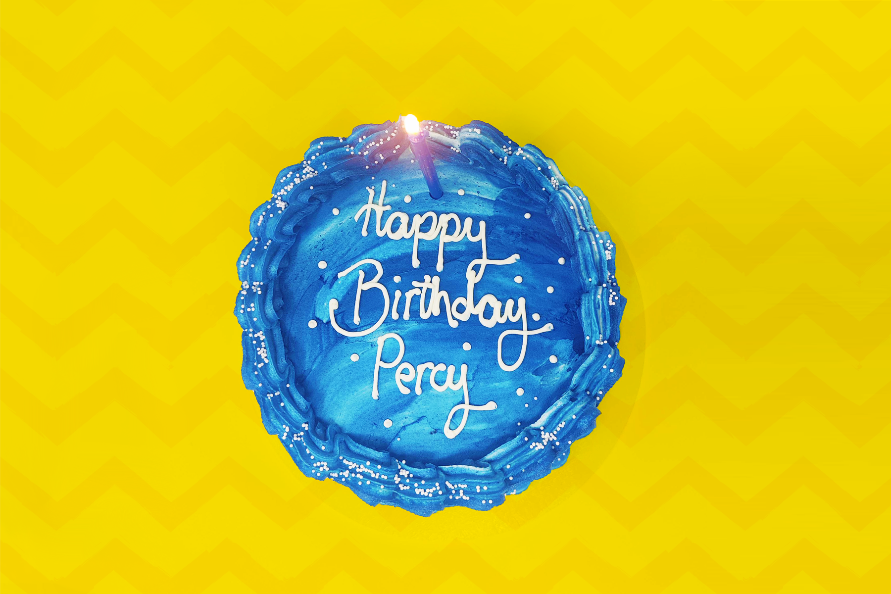 An image of a birthday cake on top of a yellow zig zag background. The cake has blue buttercream icing and has 'Happy Birthday Percy' written in white icing alongside a blue candle