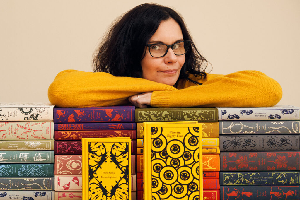 A photograph of Coralie Bickford-Smith looking over a collection of clothbound classics. Her arms are folded, she is wearing a yellow jumper. She has dark curly hair and glasses.