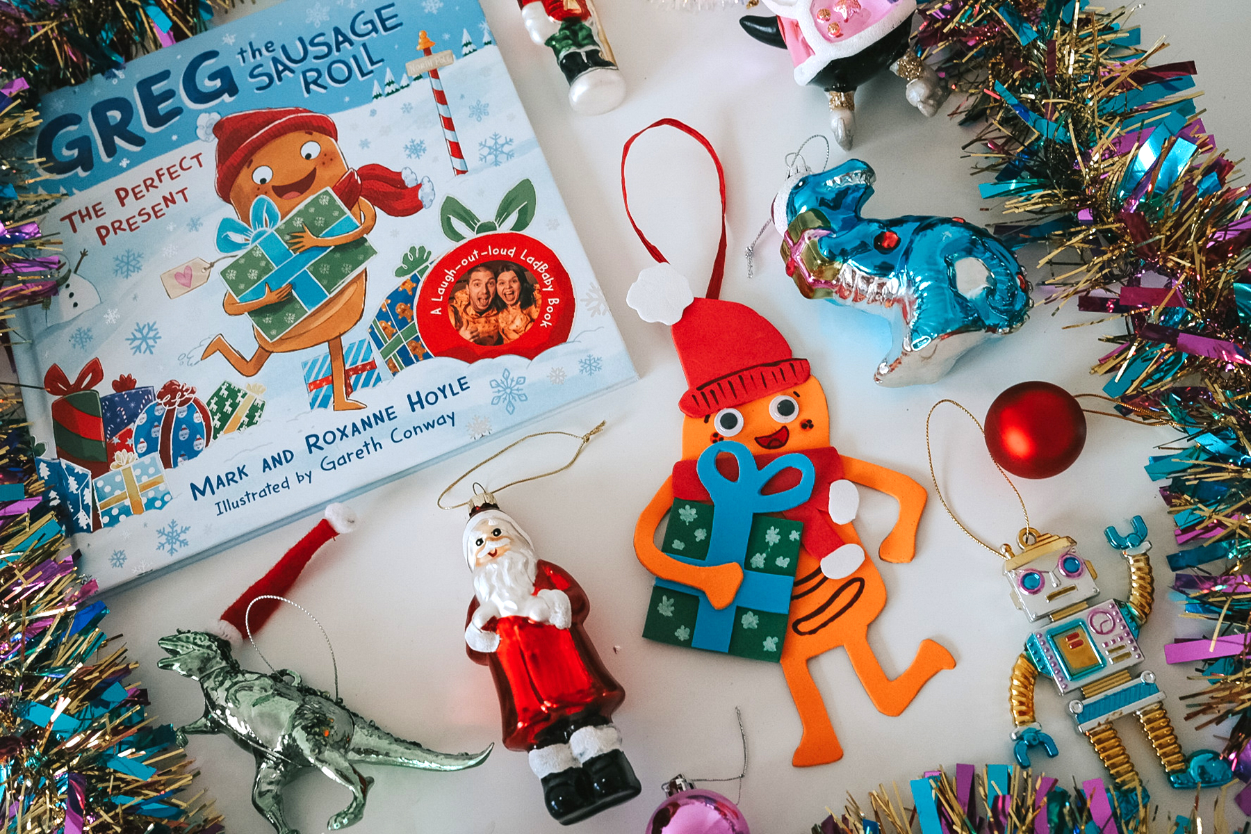 A photo of a handmade Christmas tree decoration of Greg the Sausage Roll surrounded by tinsel, baubles and other decorations, as well as a copy of the new Greg the Sausage Roll book