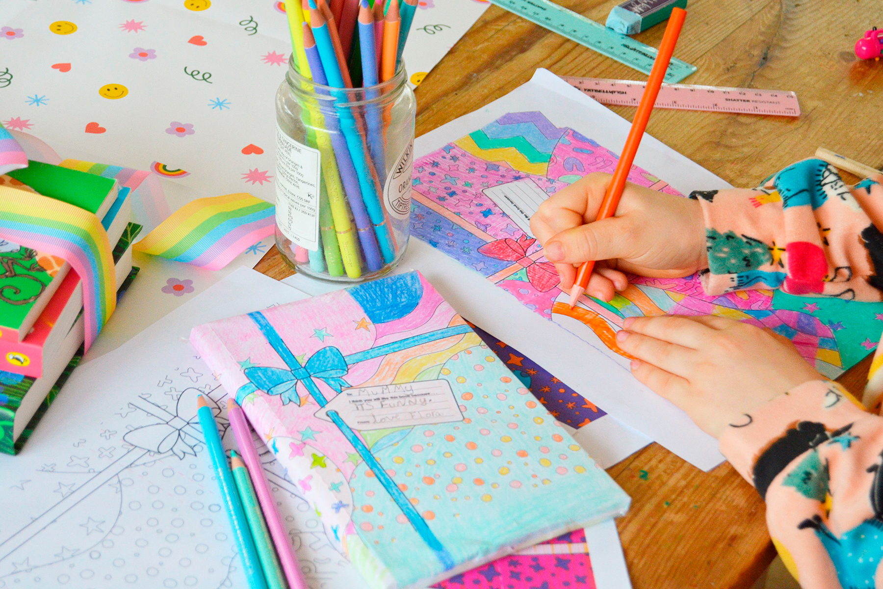 A photo of a child colouring in a book wrap on a wooden table. The child is surrounded by coloured pencils