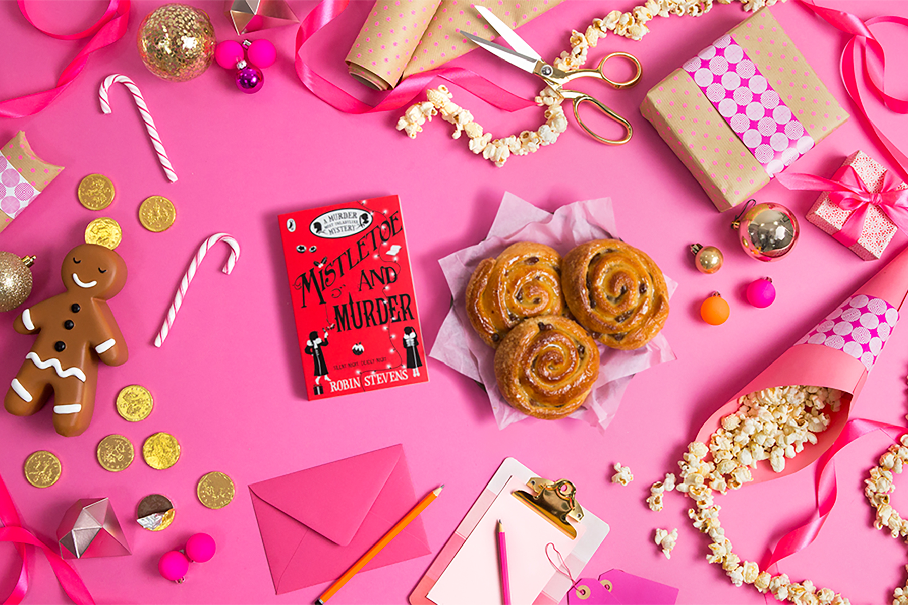 A photo of the book Mistletoe and Murder next to some Chelsea buns on a pink background surrounded by Christmas decorations
