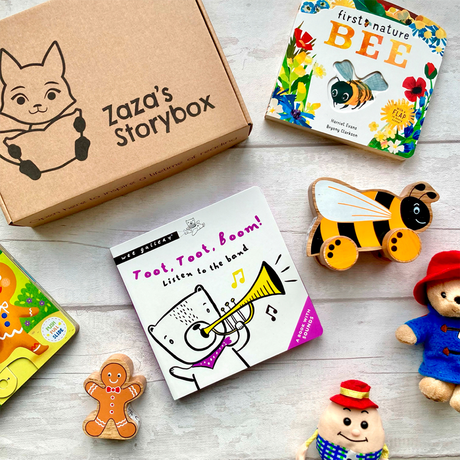 A photo of Zaza's Storybox on a white wooden background. The subscription box is made out of cardboard and is surrounded by a selection of books and some toys including Paddington Bear, Humpty Dumpty, the gingerbread man and a bee.