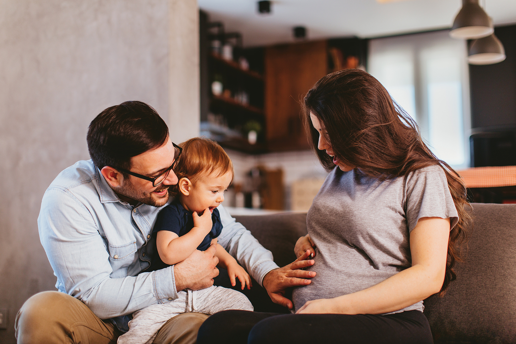 An image of two parents, mother and father, sitting with their toddler on a sofa. The mother is pregnant and talking to her child about the arrival of a new baby