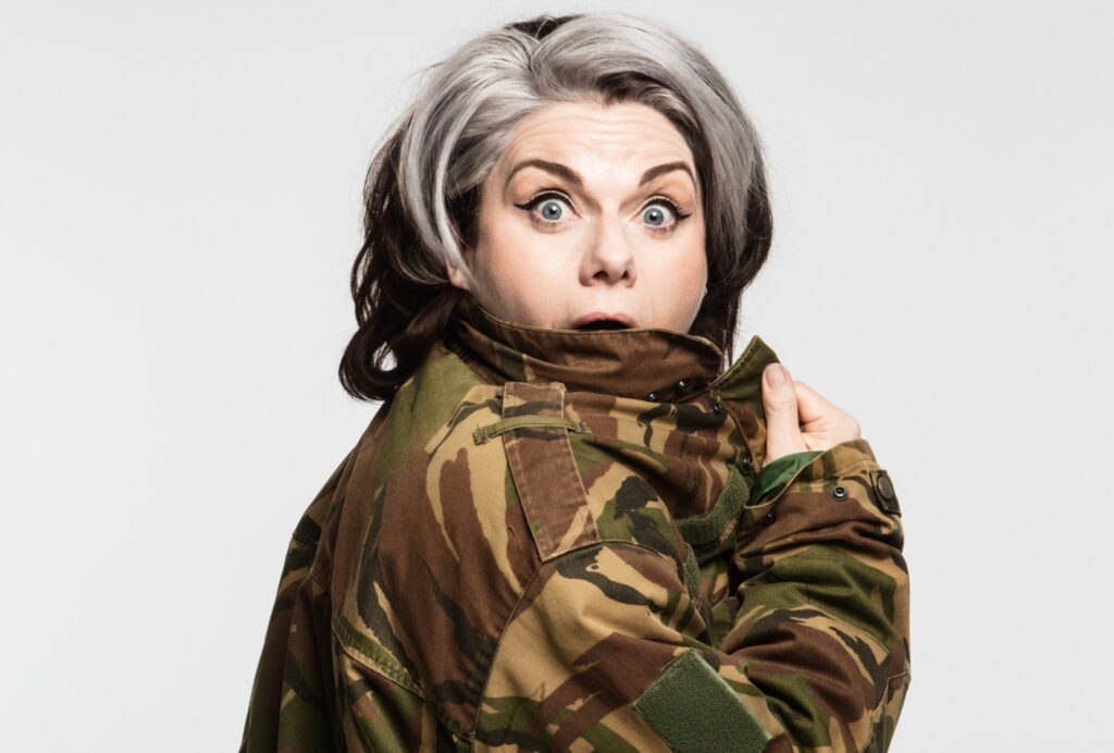 Photo of Caitlin Moran, Caitlin stands side on, against a white back ground, looking to the camera with a surprised expression, wearing a camouflage jacket.