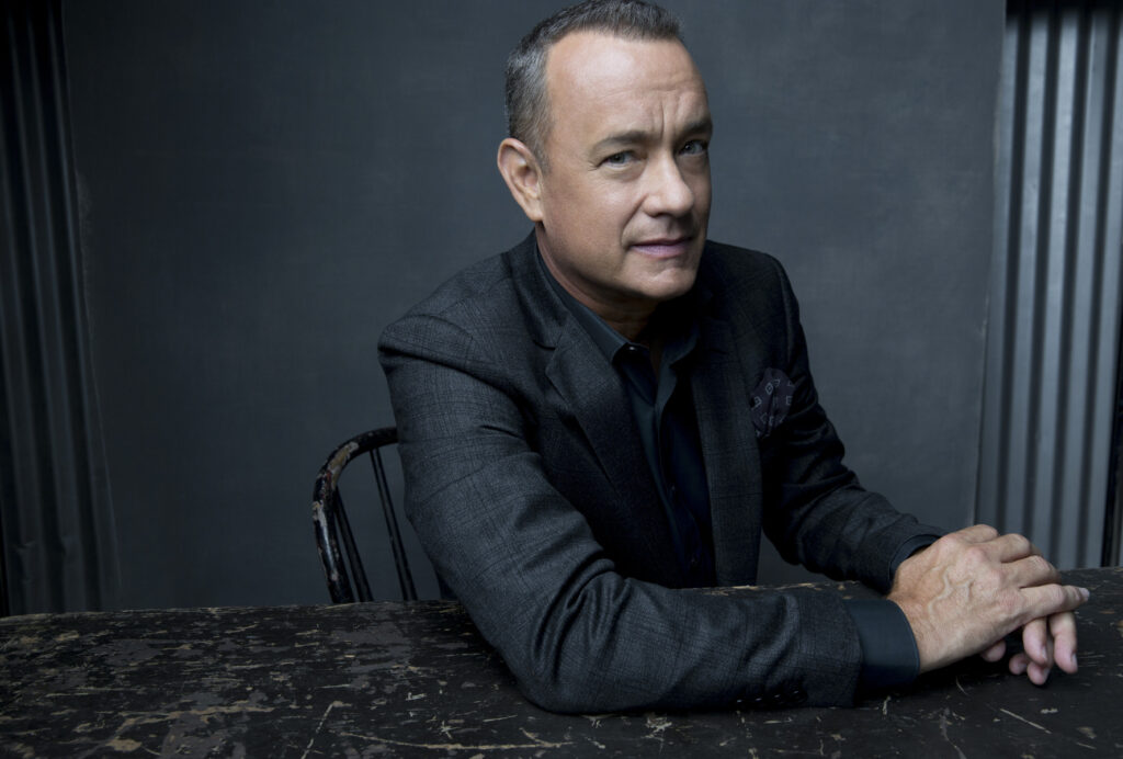 Photo of Tom Hanks, Tom sits at a table in front of a black backdrop wearing a black suit.
