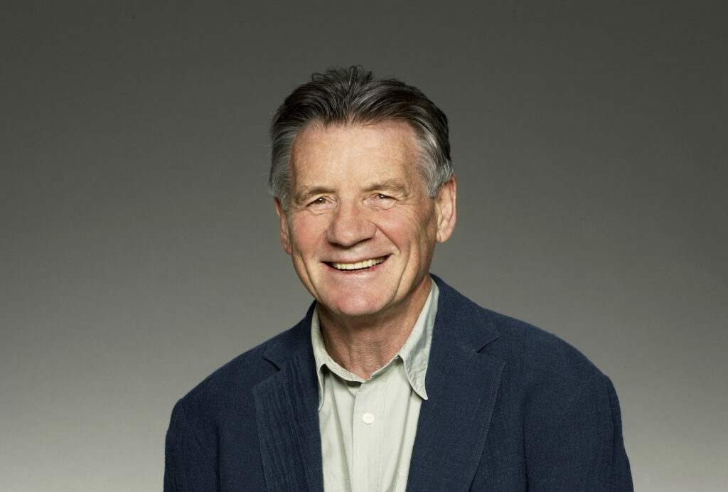 Photo of Michael Palin, Michael sits in front of a grey backdrop wearing a light green shirt and navy blue jacket.