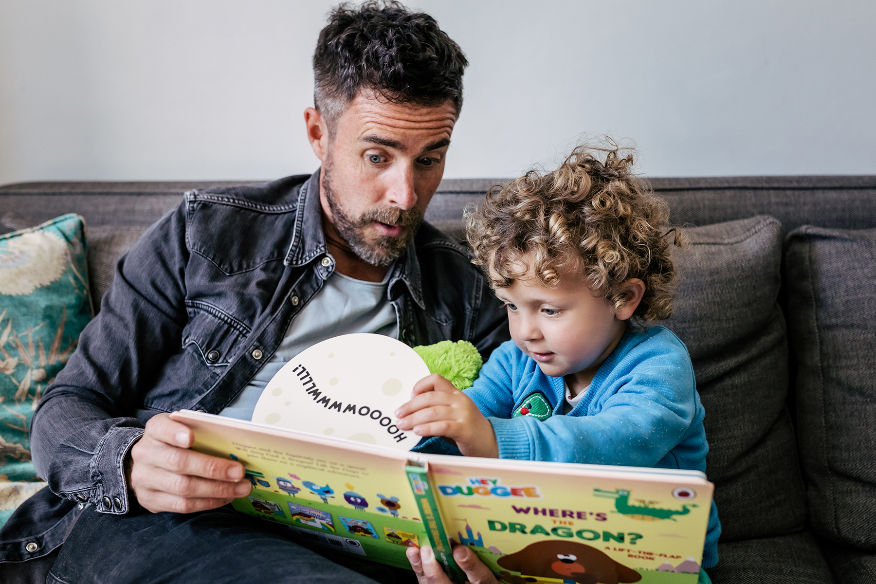 An image of a father reading a Hey Duggee book with his son. The little boy is lifting the flap on the page and his dad is making a surprised face.