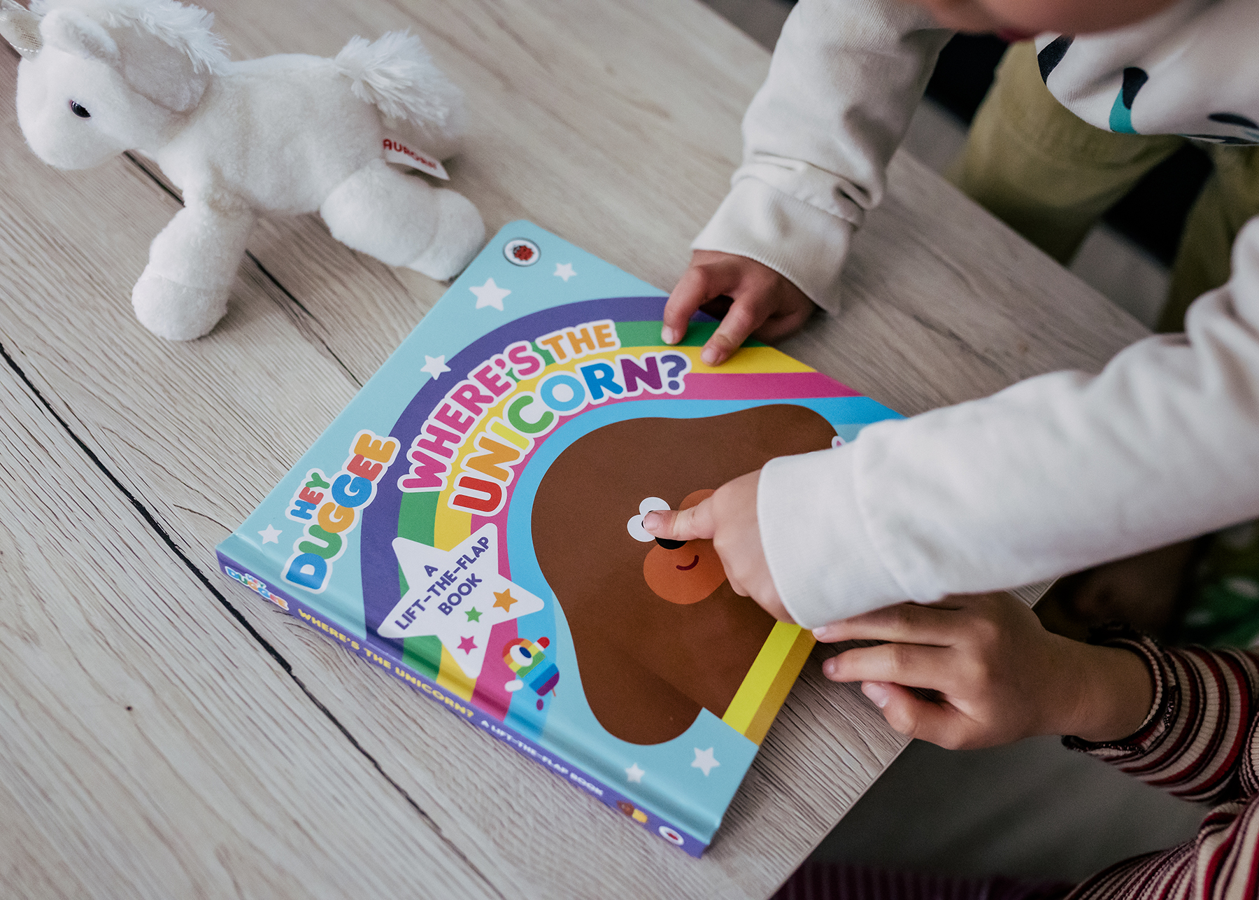 A photo of a Hey Duggee boo. There are two pairs of hands of children. One pair is pointing to Duggee's eyes on the front cover.