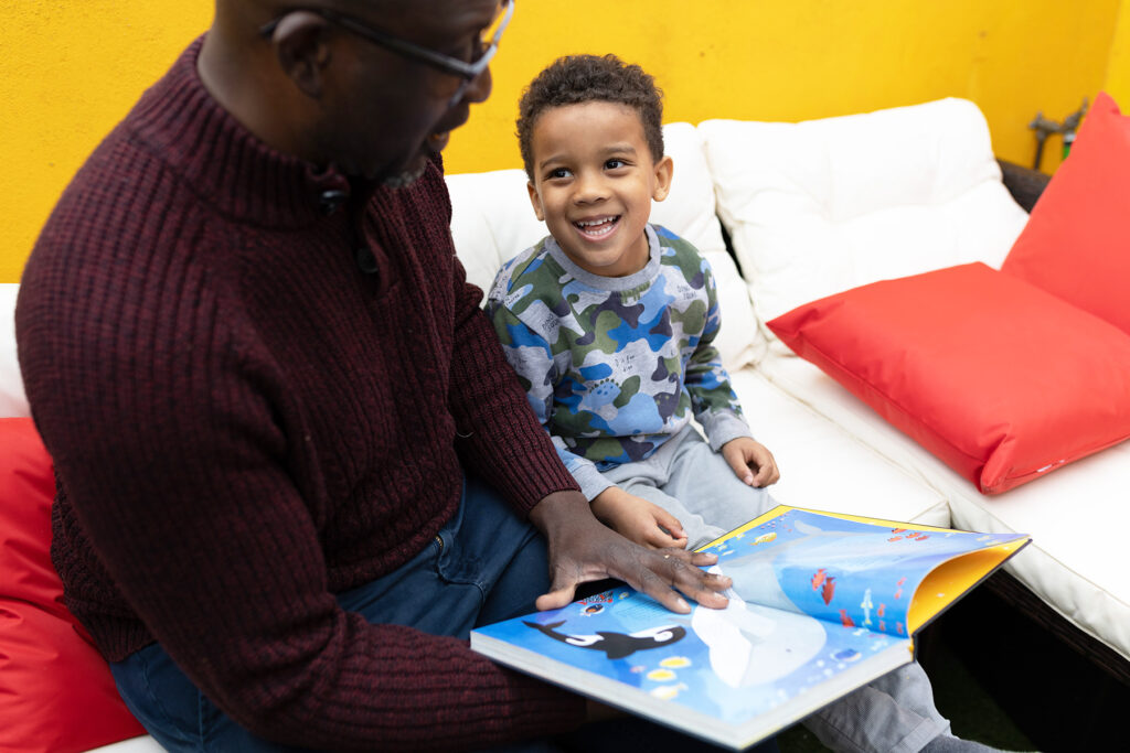 A photo of a young boy and his father reading together. The young boy is looking up at his father a smiling.