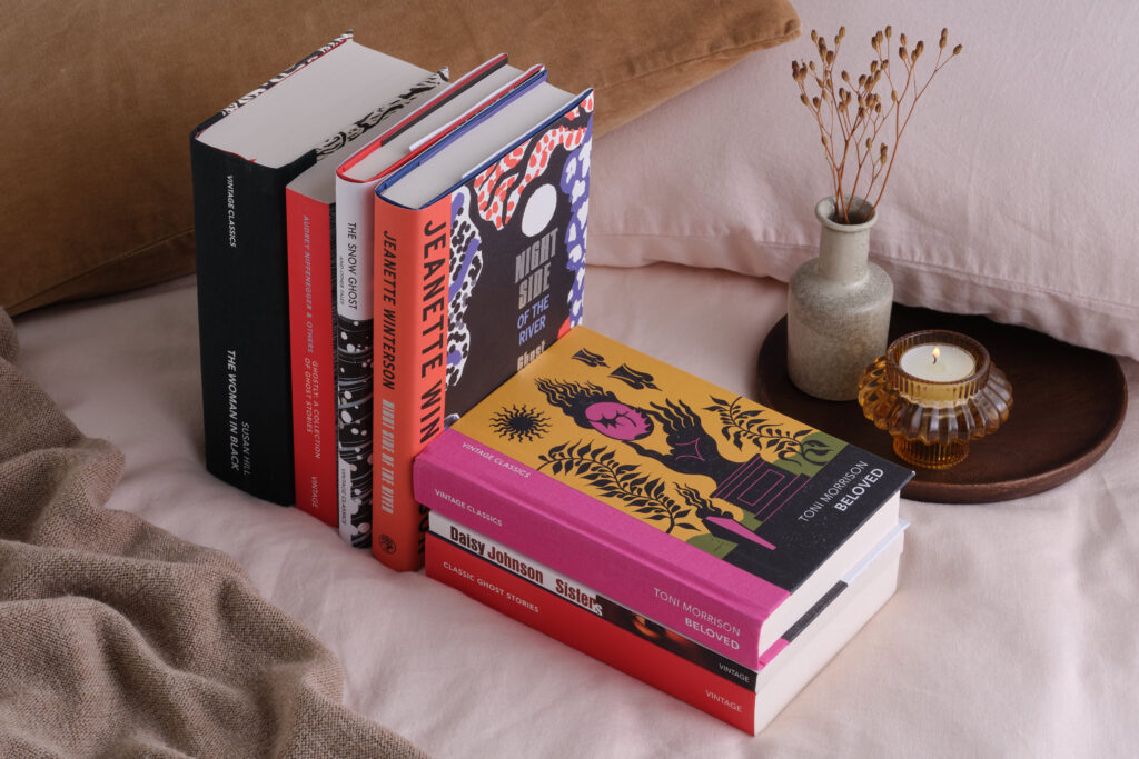 A stack of halloween books on a bed beside a candle and a vase of dried flowers.